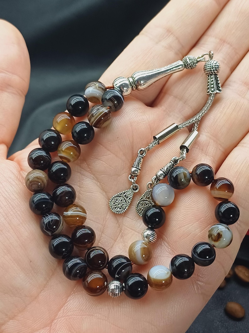 May include: A prayer bead set featuring alternating black, brown, and white agate beads. The beads are strung on a silver chain with decorative silver accents. The beads are held in a hand.