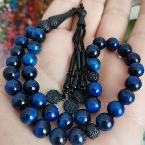 May include: A string of prayer beads featuring polished blue tiger eye stones with a chatoyant finish. The beads are connected by black cord and accented with black metal components, including a decorative tassel end and three small circular charms.