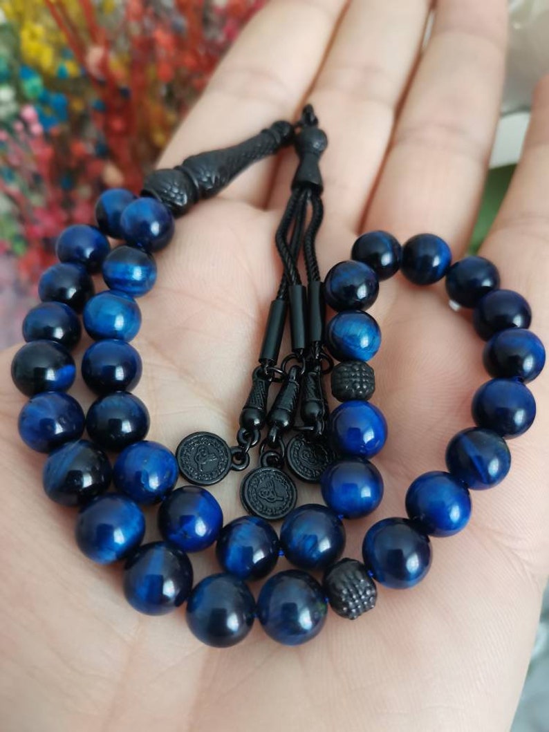 May include: A string of prayer beads featuring polished blue tiger eye stones with a chatoyant finish. The beads are connected by black cord and accented with black metal components, including a decorative tassel end and three small circular charms.