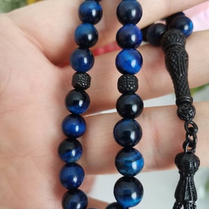 May include: A prayer bead strand featuring polished blue tiger eye gemstone spheres arranged in two parallel rows. The beads are accented with small, textured black metal spacers and a decorative black metal tassel component at the end.