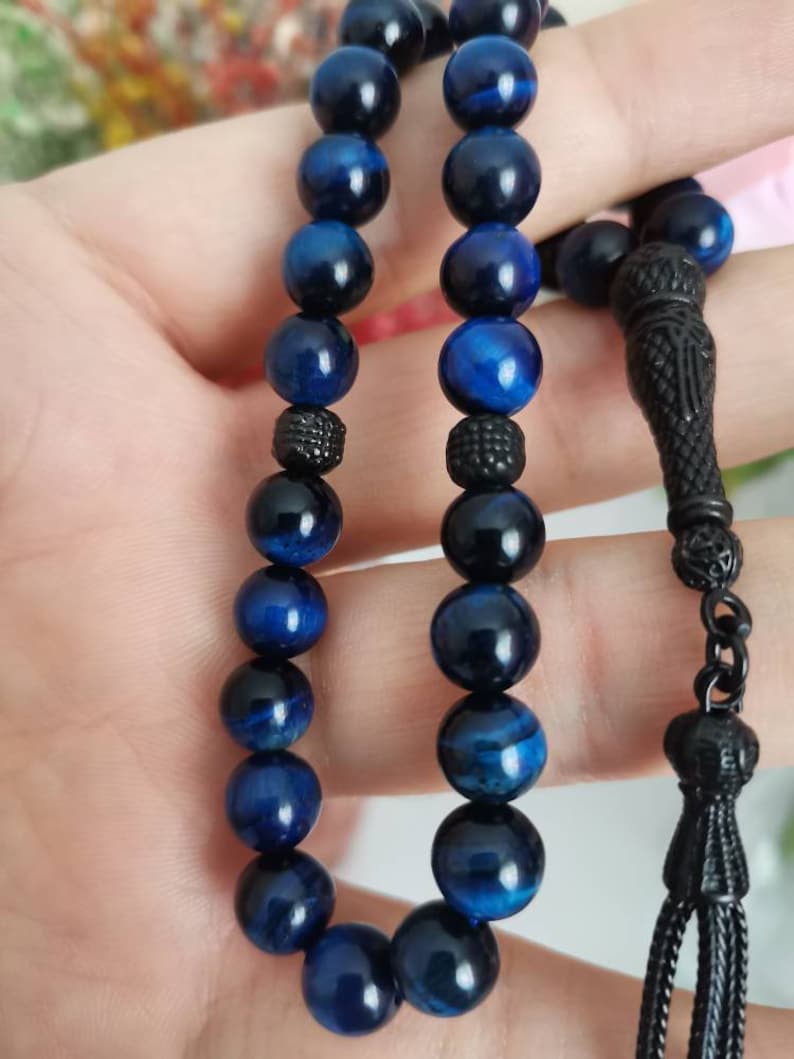 May include: A prayer bead strand featuring polished blue tiger eye gemstone spheres arranged in two parallel rows. The beads are accented with small, textured black metal spacers and a decorative black metal tassel component at the end.