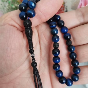 May include: A prayer bead strand featuring polished blue tiger eye gemstone beads interspersed with small, textured black spherical spacers. The strand terminates in an elongated, carved black metal pendant piece with a three-strand tassel.