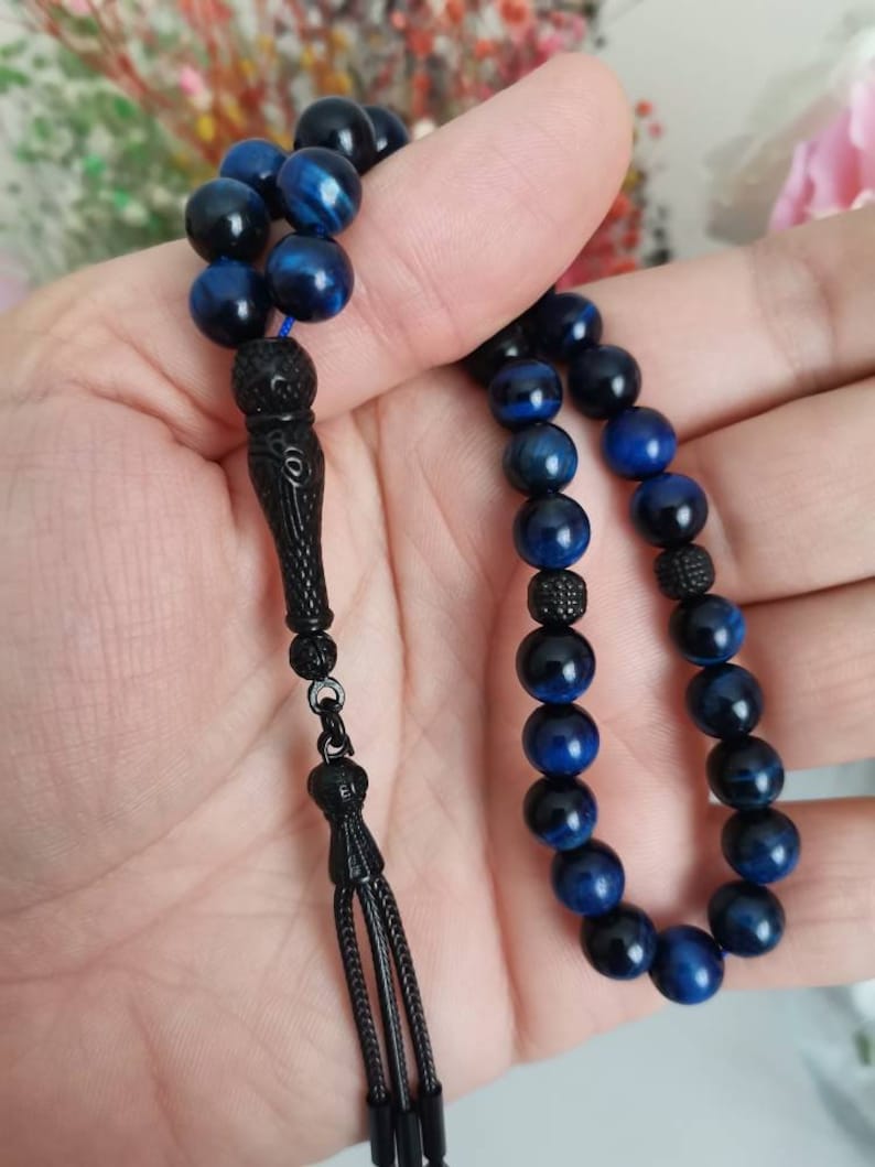 May include: A prayer bead strand featuring polished blue tiger eye gemstone beads interspersed with small, textured black spherical spacers. The strand terminates in an elongated, carved black metal pendant piece with a three-strand tassel.