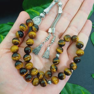 May include: A prayer bead bracelet featuring tiger eye gemstone beads. The beads are a mix of brown and gold, strung together with a silver-colored chain and decorative elements. The bracelet includes two silver tassels.