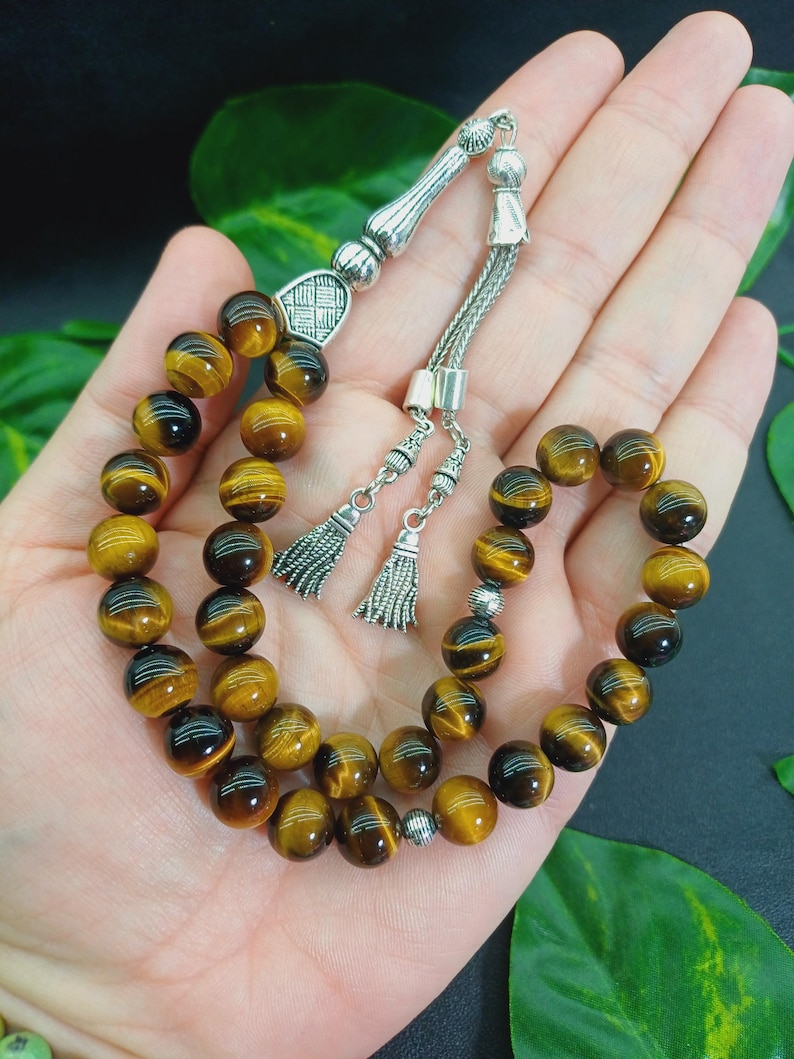 May include: A prayer bead bracelet featuring tiger eye gemstone beads. The beads are a mix of brown and gold, strung together with a silver-colored chain and decorative elements. The bracelet includes two silver tassels.