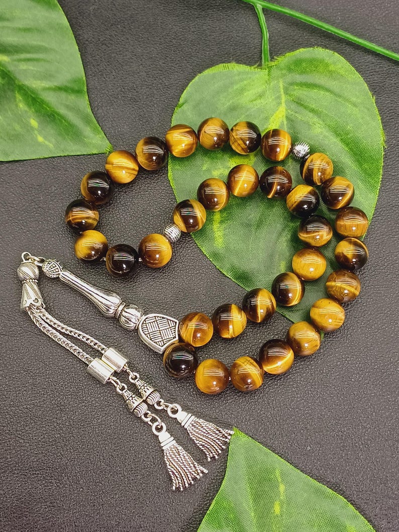 May include: A prayer bead set featuring round, tiger's eye gemstone beads in shades of brown and gold. The beads are strung together with a silver-colored metal chain and decorative elements, including tassels.