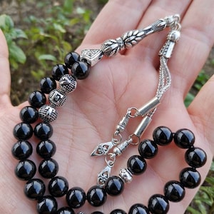 Può includere: Una collana di perline di preghiera nere con accenti in metallo color argento. Le perline sono rotonde e lucide, e i pezzi di metallo presentano disegni intricati. La collana è tenuta in una mano su uno sfondo verde sfocato.