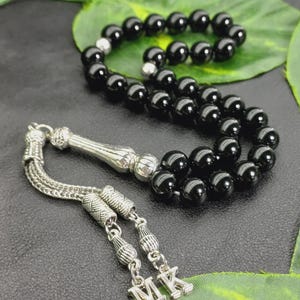 Peut inclure: Un chapelet noir avec des détails argentés. Les perles sont rondes et brillantes, reliées par une chaîne argentée. Le pompon affiche les initiales "MK" en argent. Le fond est sombre avec des accents de feuilles vertes.