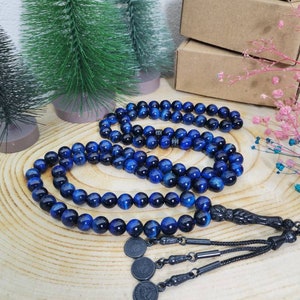 May include: A string of prayer beads featuring polished blue tiger eye gemstone spheres. The beads are connected by a black metal tassel assembly that includes three small circular charms with embossed details, ending in a black cord.