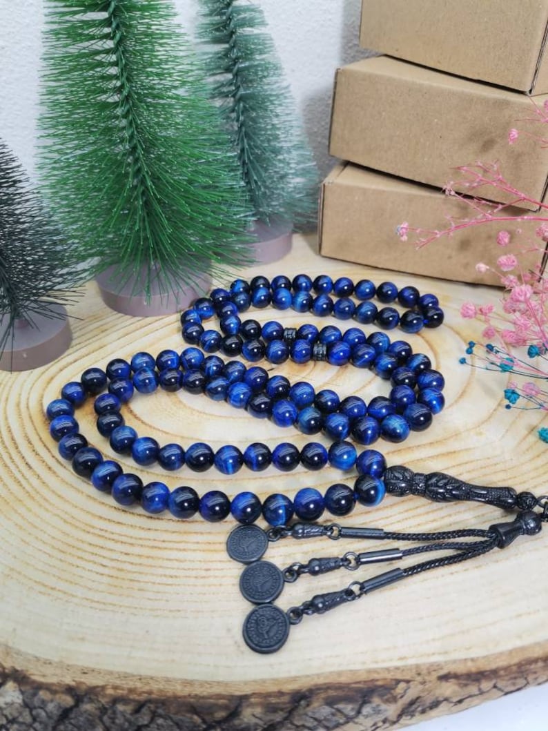 May include: A string of prayer beads featuring polished blue tiger eye gemstone spheres. The beads are connected by a black metal tassel assembly that includes three small circular charms with embossed details, ending in a black cord.
