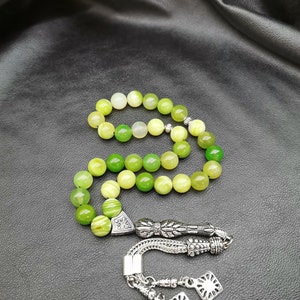 Pu&ograve; includere: Una collana di grani di preghiera verdi e gialli con elementi metallici argentati ornati. Le perle sono rotonde e variano nelle tonalit&agrave; di verde e giallo. La metalleria include motivi decorativi e un nappa.