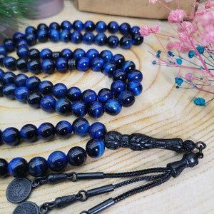 May include: A prayer bead strand featuring polished blue tiger eye gemstone beads with a chatoyant finish. The strand includes a black carved focal bead, black metal connectors, and three small circular black metal charms with embossed designs.
