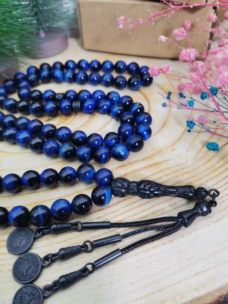 May include: A prayer bead strand featuring polished blue tiger eye gemstone beads with a chatoyant finish. The strand includes a black carved focal bead, black metal connectors, and three small circular black metal charms with embossed designs.