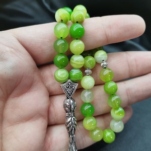 Pu&ograve; includere: Un rosario con grandi perline rotonde verdi in varie tonalit&agrave;. Le perline sono infilate insieme con un divisore e un nappa in metallo color argento. Le perline sono tenute in una mano su uno sfondo scuro.