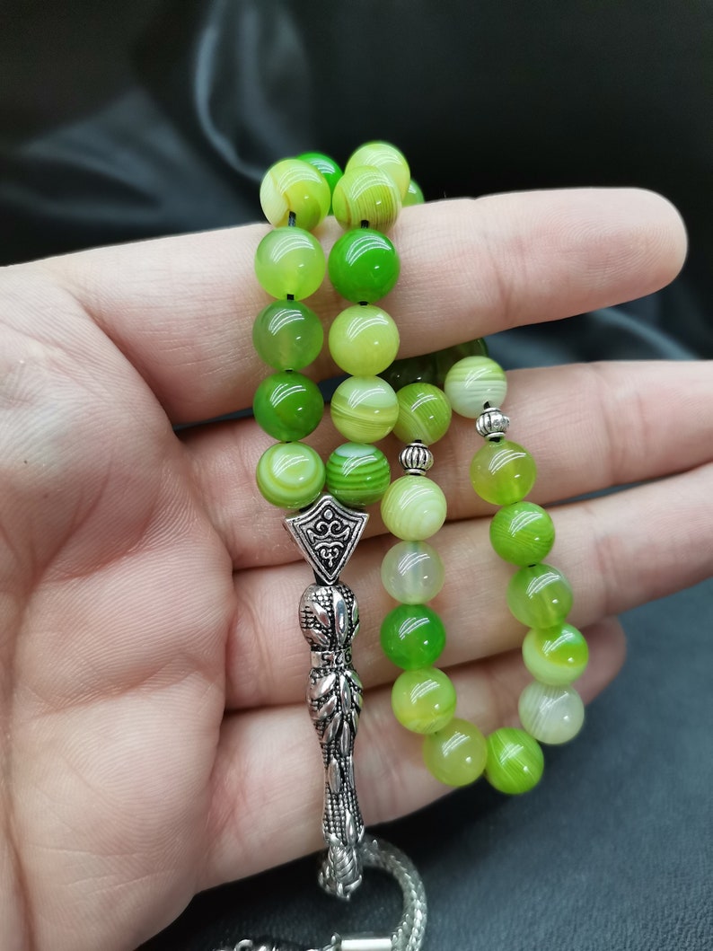 Pu&ograve; includere: Un rosario con grandi perline rotonde verdi in varie tonalit&agrave;. Le perline sono infilate insieme con un divisore e un nappa in metallo color argento. Le perline sono tenute in una mano su uno sfondo scuro.