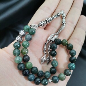 May include: A strand of prayer beads with green and silver accents. The beads are a mix of dark and light green, with a silver tassel and decorative silver pieces. The beads are held in an open hand.