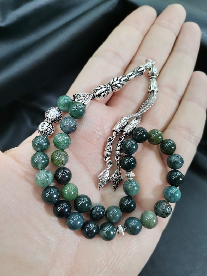 May include: A strand of prayer beads with green and silver accents. The beads are a mix of dark and light green, with a silver tassel and decorative silver pieces. The beads are held in an open hand.
