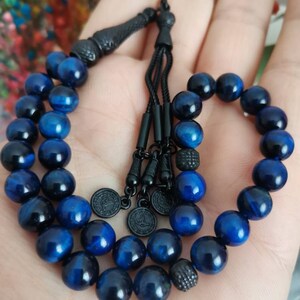May include: A prayer bead strand featuring polished blue tiger eye gemstone beads. The strand includes black metal spacers, a black tassel component, and three small circular black metal charms with engraved details, all strung on a black cord.