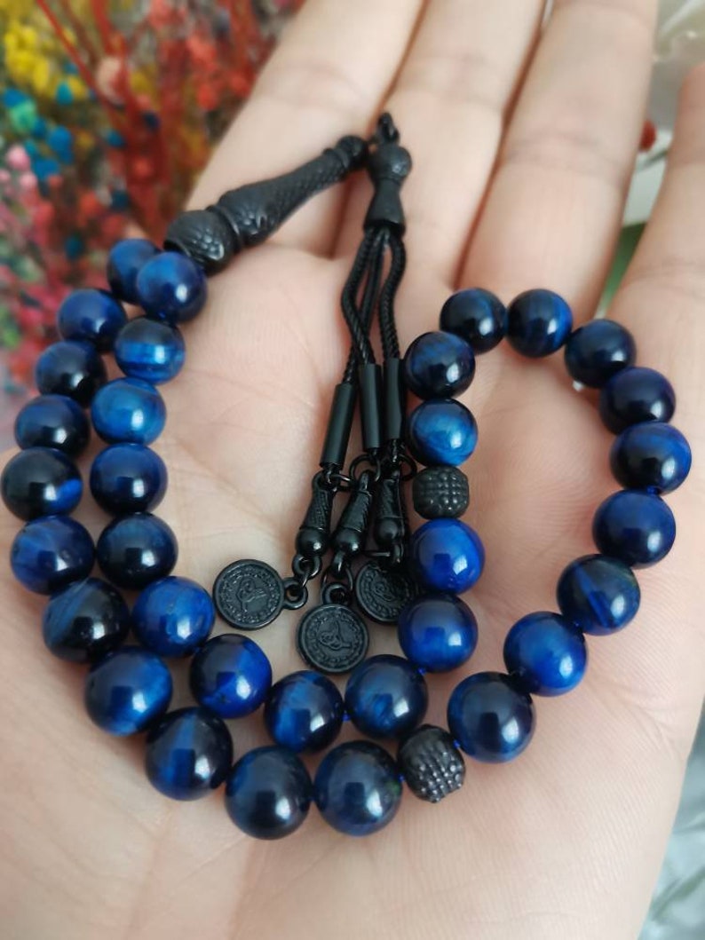 May include: A prayer bead strand featuring polished blue tiger eye gemstone beads. The strand includes black metal spacers, a black tassel component, and three small circular black metal charms with engraved details, all strung on a black cord.