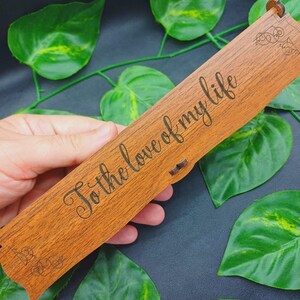 May include: A rectangular wooden box with a hinged lid featuring the laser-engraved text "To the love of my life" in a cursive script. The box has a natural brown wood grain finish and decorative scrollwork patterns etched at the corners.