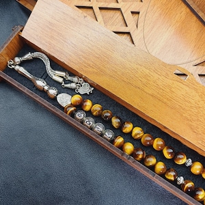 Può includere: Una scatola di legno con un design geometrico decorativo contiene una collana di perline. La collana presenta perline occhio di tigre e dettagli in metallo argentato. La scatola è aperta, rivelando la collana.