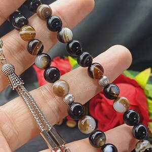 May include: A beaded necklace featuring alternating black, brown, and white striped agate beads. The beads are strung on a silver chain with decorative silver accents. The necklace is displayed against a dark background.