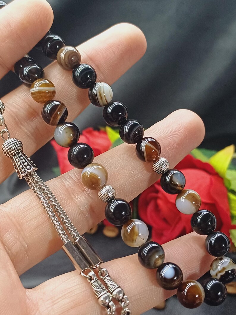 May include: A beaded necklace featuring alternating black, brown, and white striped agate beads. The beads are strung on a silver chain with decorative silver accents. The necklace is displayed against a dark background.