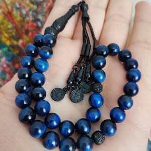 May include: A prayer bead strand featuring polished blue tiger eye gemstone beads. The strand includes black metal spacers, a black tassel component, and three small circular black metal charms with engraved details, all strung on a black cord.