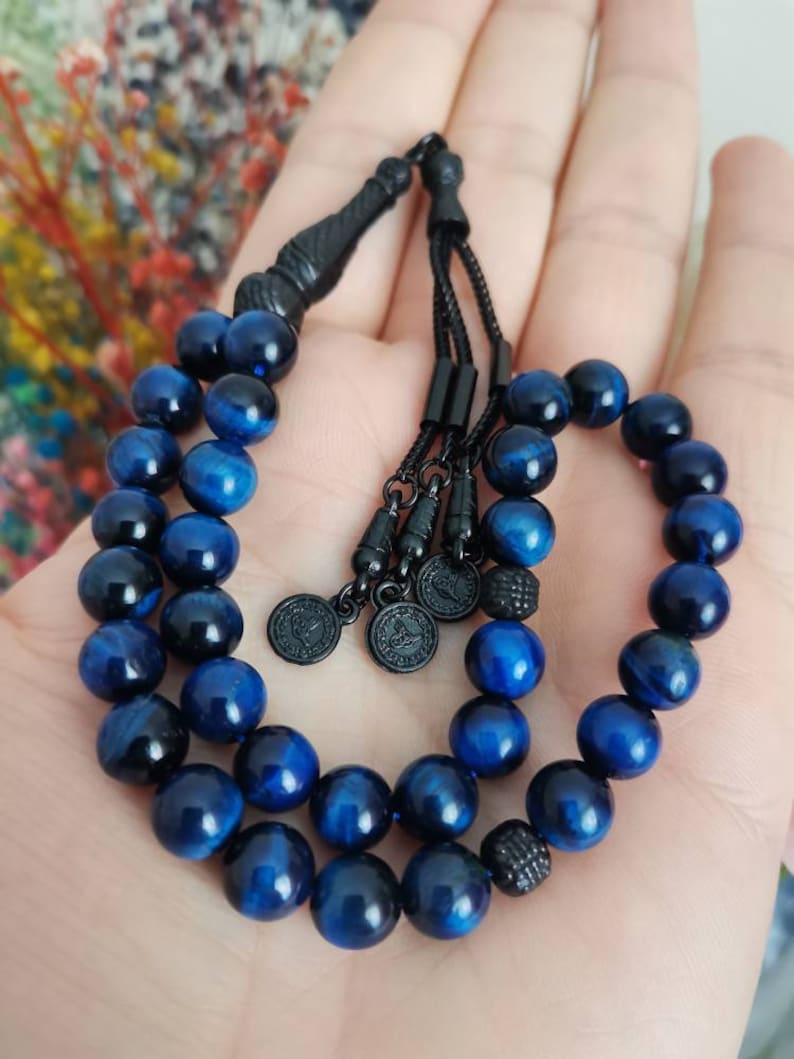 May include: A prayer bead strand featuring polished blue tiger eye gemstone beads. The strand includes black metal spacers, a black tassel component, and three small circular black metal charms with engraved details, all strung on a black cord.