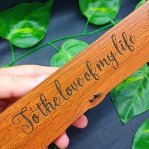 May include: A rectangular wooden box with the phrase "To the love of my life" engraved in a cursive font. The box is a warm brown color and has a small latch. Green leaves are in the background.