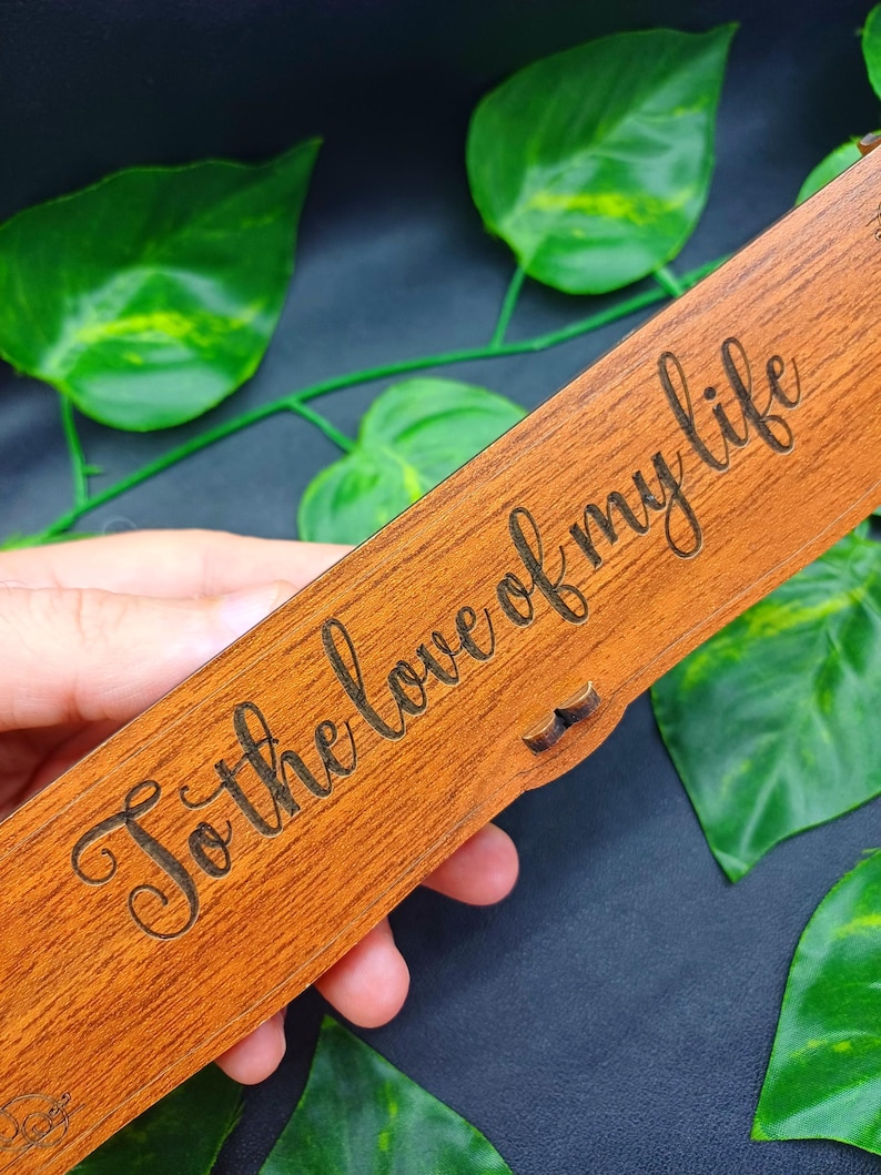 May include: A rectangular wooden box with the phrase "To the love of my life" engraved in a cursive font. The box is a warm brown color and has a small latch. Green leaves are in the background.