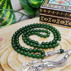 May include: A green beaded necklace with silver accents, resting on a wooden surface. The beads are a deep emerald green, and the necklace has a decorative silver tassel. A small, decorated wooden box is in the background.