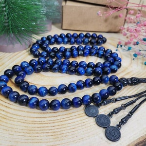 May include: A string of prayer beads featuring polished blue tiger eye stones with a chatoyant finish. The beads are connected by a black cord, ending in a black metal tassel component with three circular charms, each embossed with a detailed emblem.