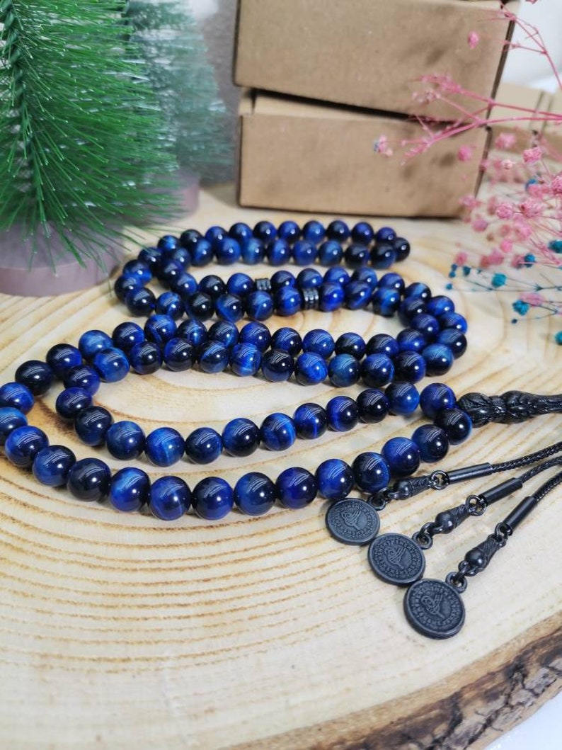May include: A string of prayer beads featuring polished blue tiger eye stones with a chatoyant finish. The beads are connected by a black cord, ending in a black metal tassel component with three circular charms, each embossed with a detailed emblem.
