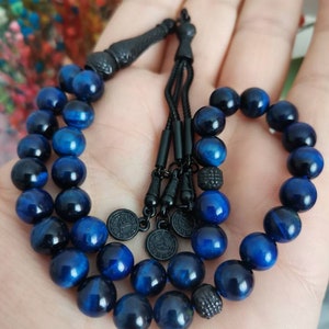 May include: A prayer bead strand featuring polished, deep blue tiger eye gemstone beads. The strand includes black metal spacers, a black tassel component, and three small circular black metal charms with engraved details on the end of the cord.