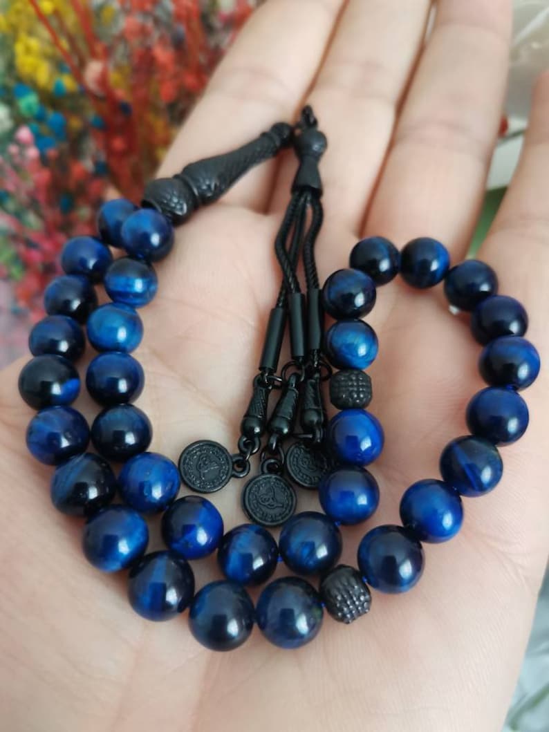 May include: A prayer bead strand featuring polished, deep blue tiger eye gemstone beads. The strand includes black metal spacers, a black tassel component, and three small circular black metal charms with engraved details on the end of the cord.