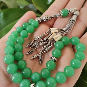 May include: A green beaded prayer rosary with silver-coloured metal accents. The beads are a vibrant, solid green colour. The rosary features decorative metal feather charms and a tassel.