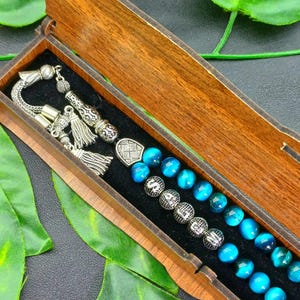 Puede incluir: Una caja de madera con tapa con bisagras contiene un juego de cuentas de oración. Las cuentas son de un azul vibrante con efecto veteado. Acentos y borlas de metal plateado adornan los extremos de la hebra. La caja es de madera de tonos cálidos.