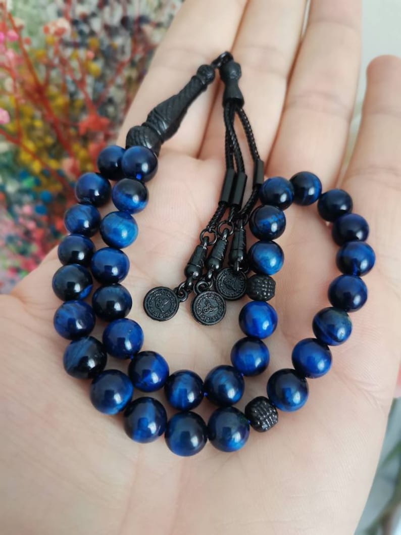 May include: A prayer bead strand featuring polished, spherical blue tiger eye beads with a chatoyant luster. The strand includes black metal spacers, a black tassel connector, and three small circular black metal charms with embossed designs.