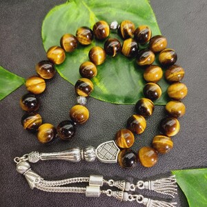 May include: A strand of prayer beads with round, tiger's eye beads in shades of brown and gold. The beads are strung together with a silver-colored metal divider and tassel. The background includes a green leaf and a dark surface.