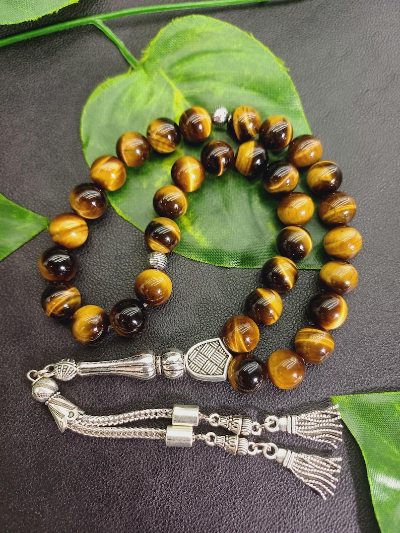May include: A strand of prayer beads with round, tiger's eye beads in shades of brown and gold. The beads are strung together with a silver-colored metal divider and tassel. The background includes a green leaf and a dark surface.