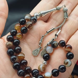 May include: A beaded bracelet featuring alternating black, brown, and cream-colored agate beads. The bracelet has silver-colored metal accents and a decorative tassel. The beads are approximately 0.3 inches in diameter.