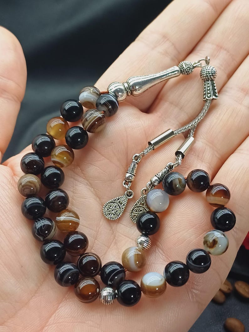 May include: A beaded bracelet featuring alternating black, brown, and cream-colored agate beads. The bracelet has silver-colored metal accents and a decorative tassel. The beads are approximately 0.3 inches in diameter.