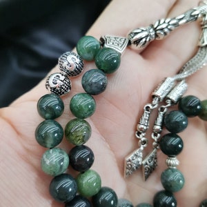 May include: A close-up of a green beaded prayer beads with silver accents. The beads are a mix of dark and light green, with a polished finish. The silver components feature intricate floral and geometric designs.