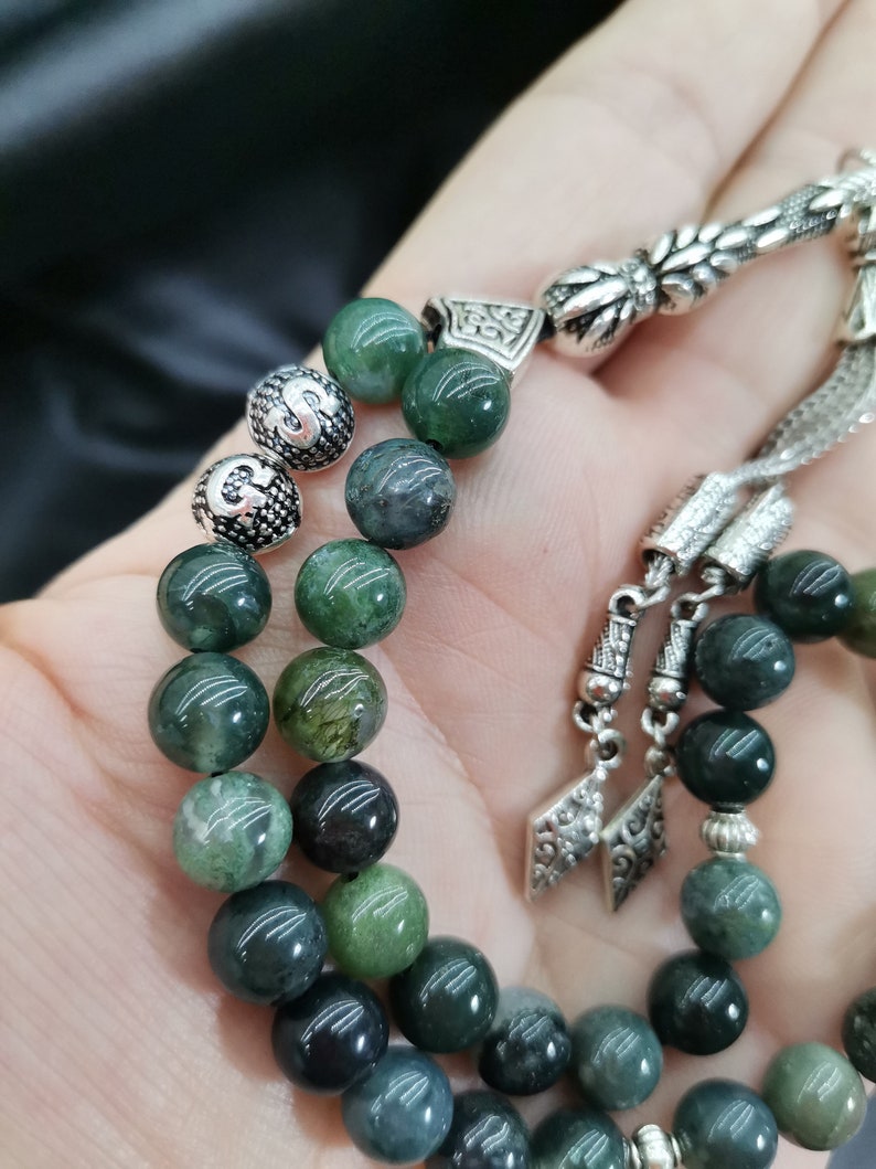 May include: A close-up of a green beaded prayer beads with silver accents. The beads are a mix of dark and light green, with a polished finish. The silver components feature intricate floral and geometric designs.