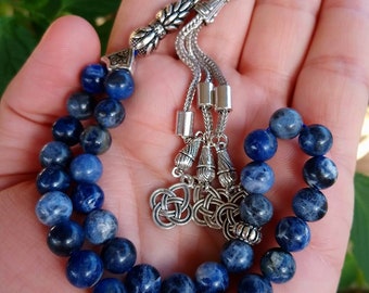 Op de afbeelding: Een gebedsketting met donkerblauwe, bolvormige kralen. De kralen zijn geregen op een zilverkleurige ketting met decoratieve metalen elementen. Het ontwerp omvat sierlijk metaalwerk aan de uiteinden en een decoratieve bedel.