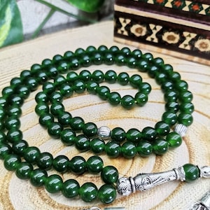 May include: A dark green beaded necklace with silver-colored accents. The beads are round and evenly sized. The necklace is draped on a wooden surface, with a decorative box in the background.