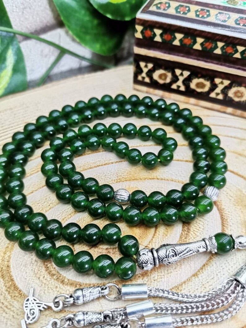 May include: A dark green beaded necklace with silver-colored accents. The beads are round and evenly sized. The necklace is draped on a wooden surface, with a decorative box in the background.