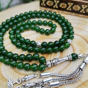 May include: A strand of dark green prayer beads, or a rosary, with a silver tassel and decorative metal accents. The beads are round and smooth, and the tassel is made of multiple strands. The background is a wooden surface.