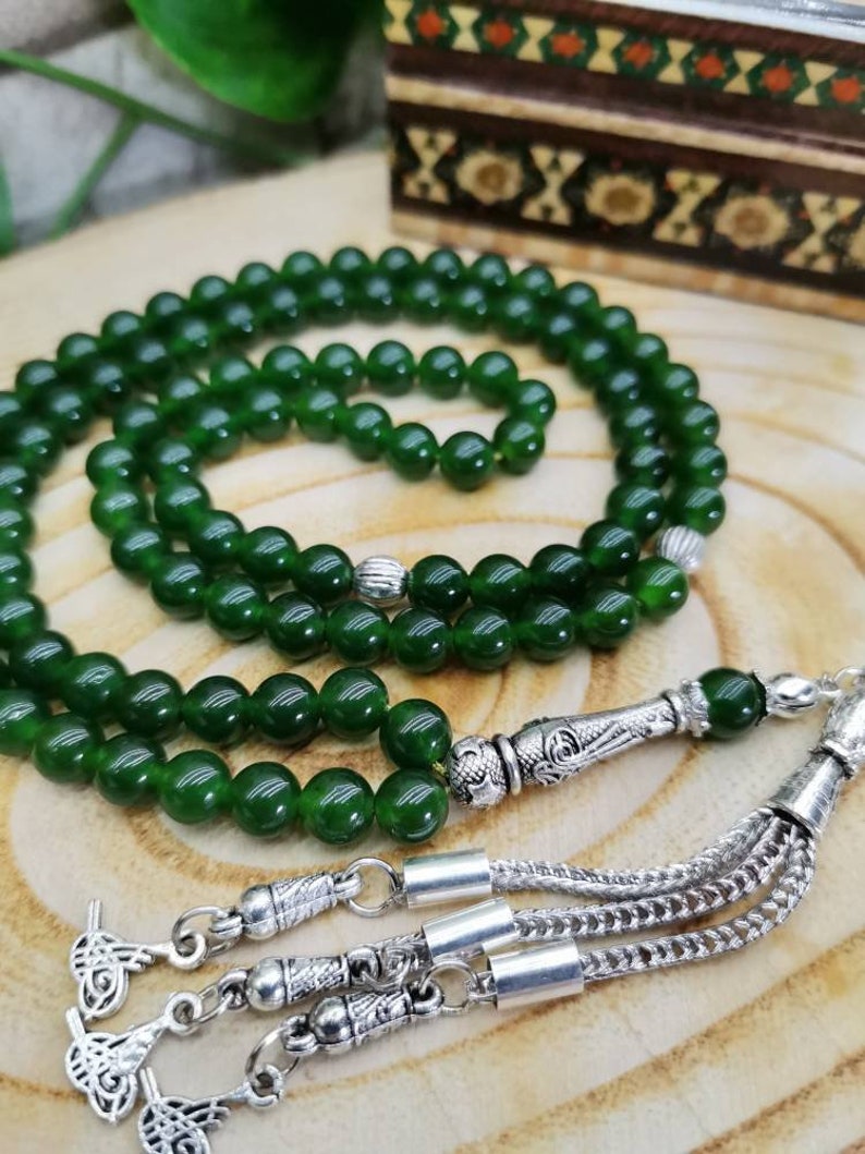 May include: A strand of dark green prayer beads, or a rosary, with a silver tassel and decorative metal accents. The beads are round and smooth, and the tassel is made of multiple strands. The background is a wooden surface.